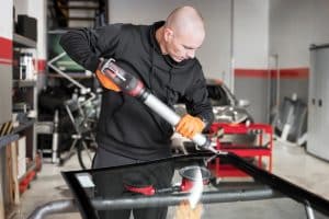 A technician in a garage applies adhesive to a new windshield using a specialised tool, preparing it for installation. This image underscores the meticulous process of windshield replacement, emphasizing the crucial steps to ensure safety before you "drive after windshield replacement."