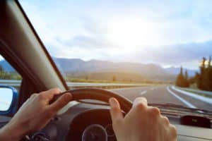A driver's hands gripping the steering wheel while driving on an open highway with mountains in the background under a bright sky. This image captures the essence of a safe and scenic drive after windshield replacement, highlighting the importance of ensuring a clear and secure windshield for optimal driving conditions.