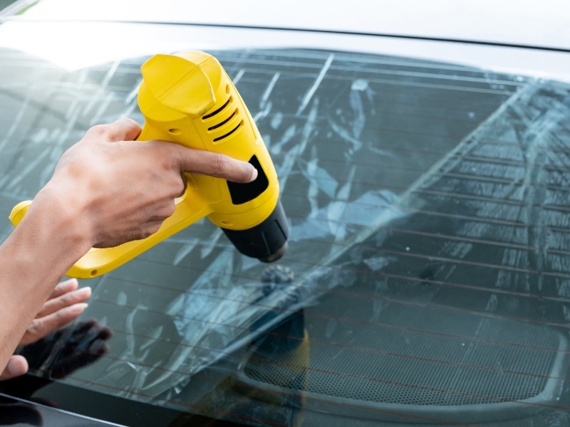 Technician using heat gun to remove film, showing factors affecting rear windscreen replacement cost